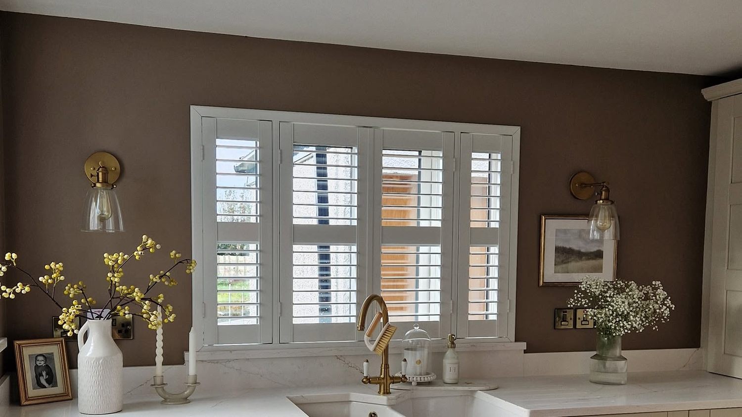 White plantation shutters fitted across a kitchen window above a belfast sink, louvres tilted to filter the daylight
